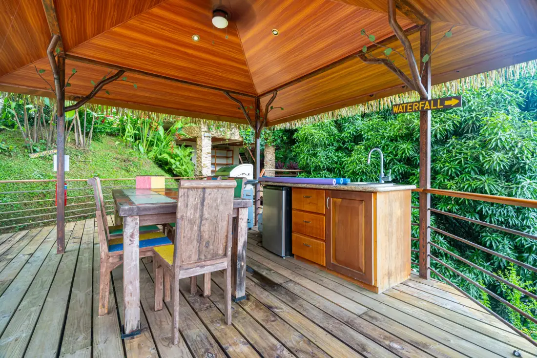 An outdoor wooden deck has a dining table and a built-in counter surrounded by greenery.