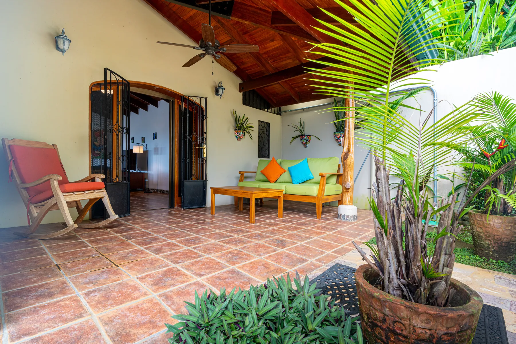 A covered patio with seating, a wooden table, and tropical plants.