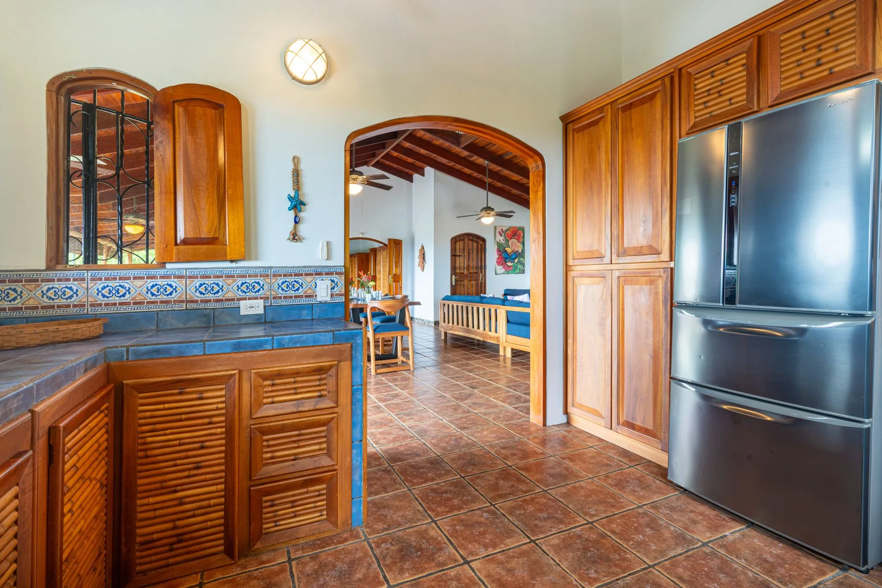 A kitchen with wooden cabinetry and a stainless steel refrigerator opens into a tiled dining area.