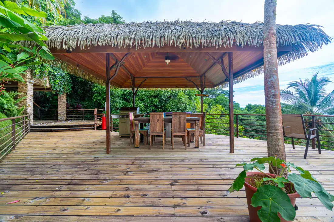 A wooden outdoor dining area sits beneath a thatched pavilion, surrounded by tropical greenery.