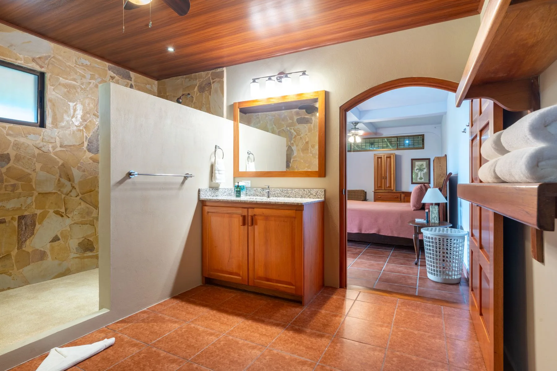 A bathroom with a walk-in shower, wooden vanity, and an open doorway to a bedroom.