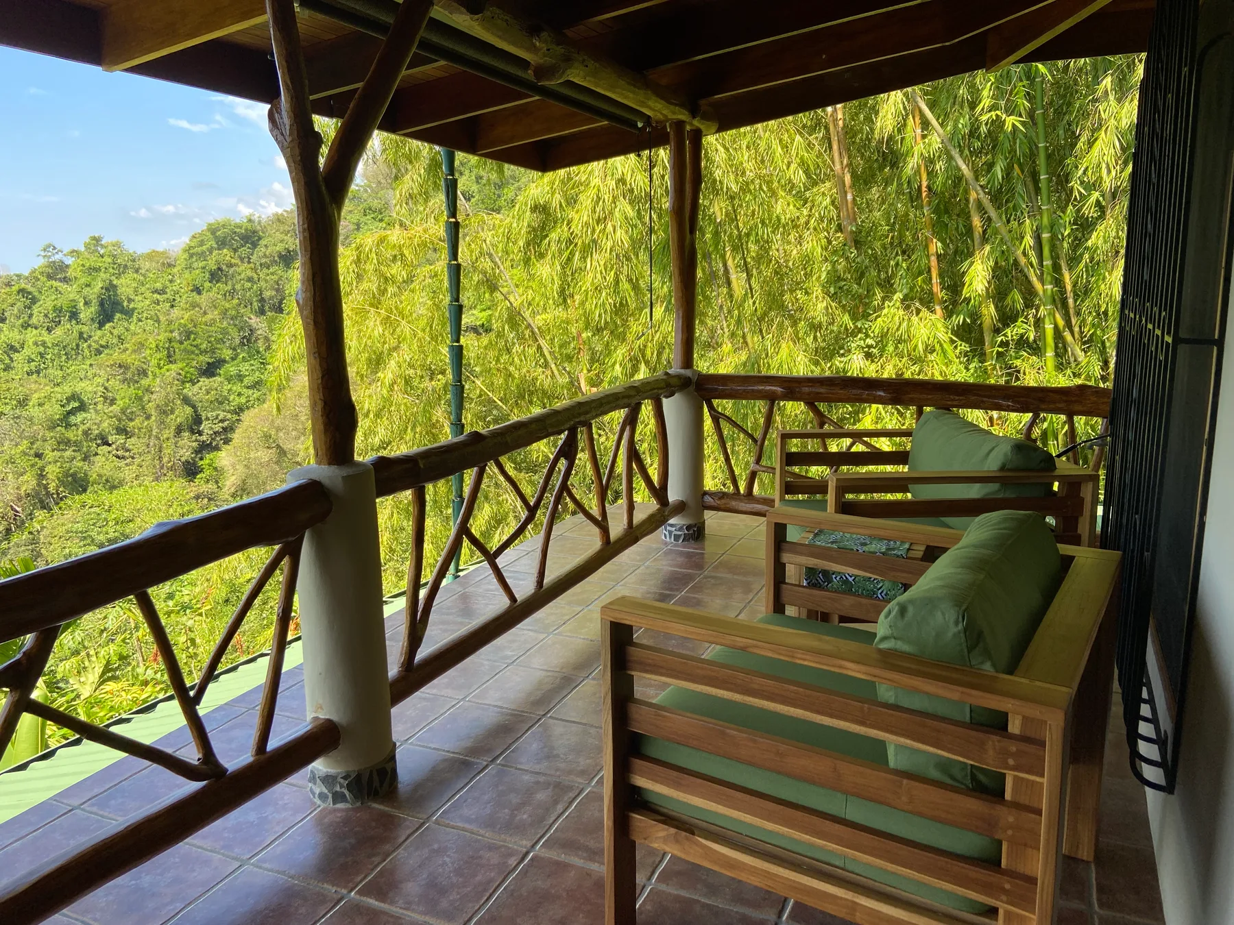 A covered wooden balcony with cushioned seating overlooks dense green vegetation.