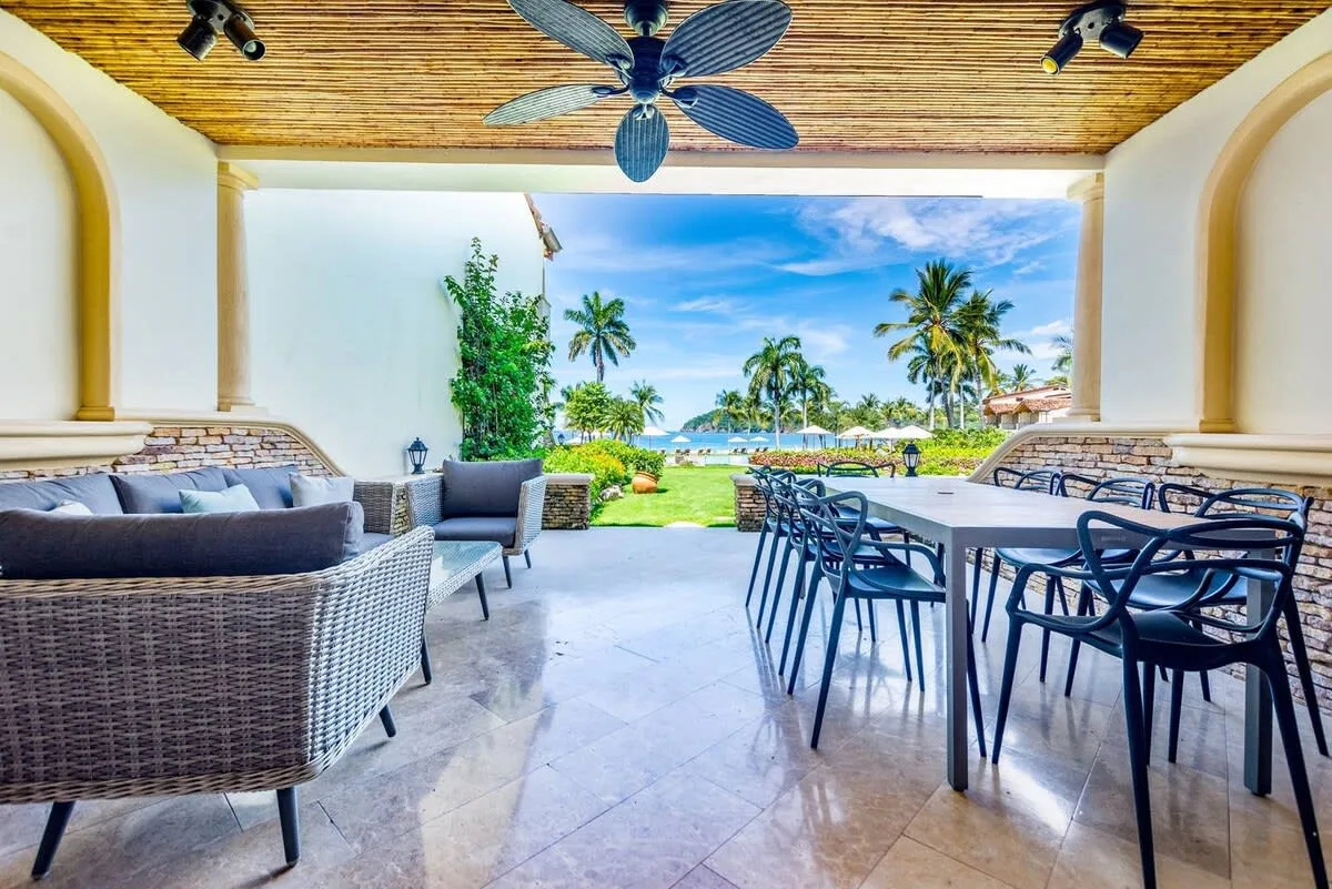 A covered outdoor dining and seating area overlooks a garden with palm trees.