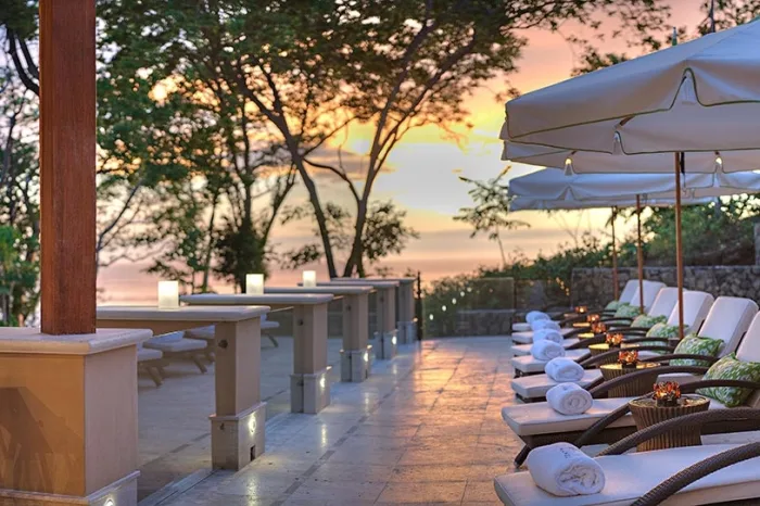 Una terraza al aire libre tiene asientos y mesas bajo grandes sombrillas al atardecer.