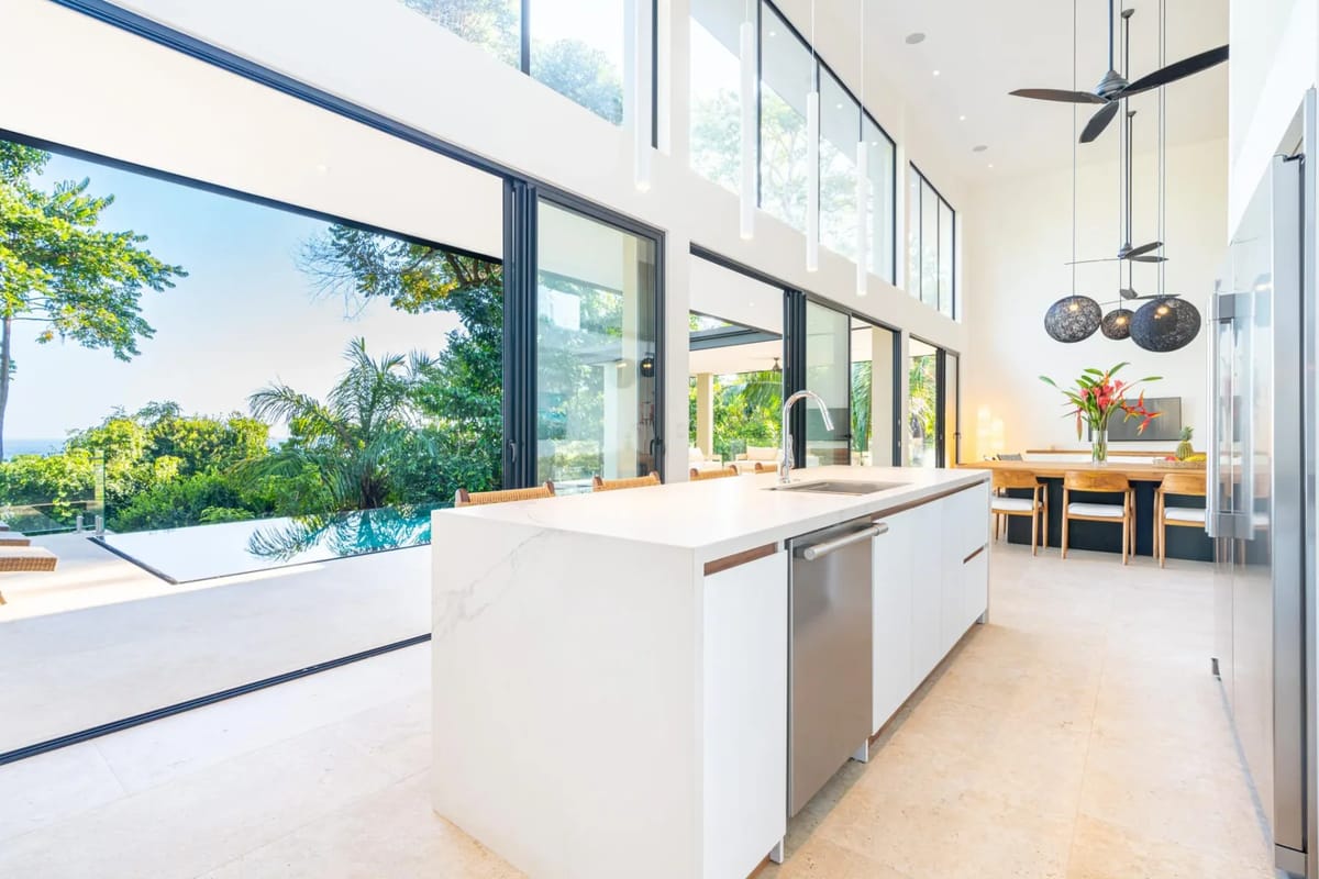 Colíbri Sur · 1. The high-ceilinged kitchen with a large island and adjacent dining area receives abundant natural light and opens through expansive glass doors to an infinity pool and tropical ocean views.