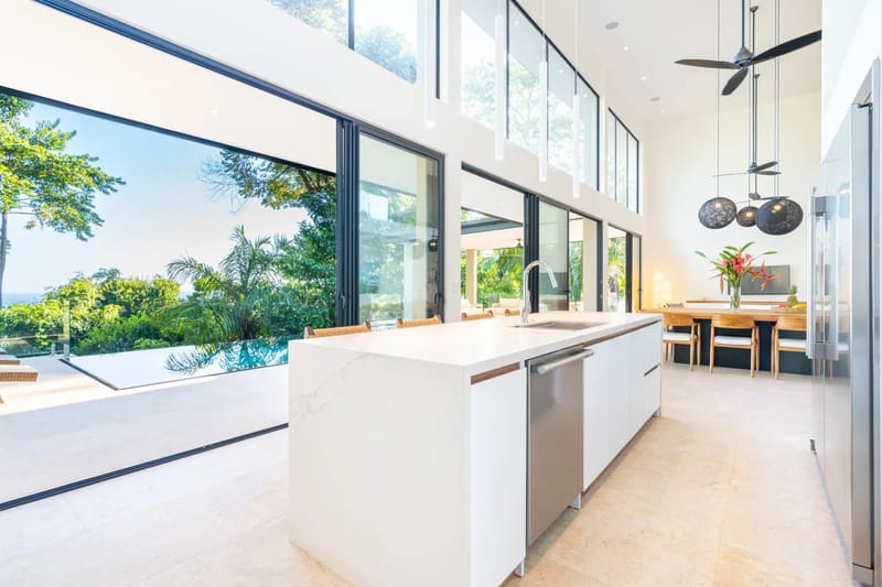 Colíbri Sur · 1. The high-ceilinged kitchen with a large island and adjacent dining area receives abundant natural light and opens through expansive glass doors to an infinity pool and tropical ocean views.