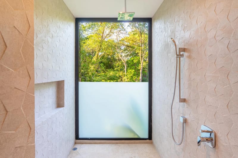 Colíbri Sur · 8. This spacious shower features textured wall tiles, a rainfall showerhead, and a large window with a frosted lower half offering private views of tropical trees.
