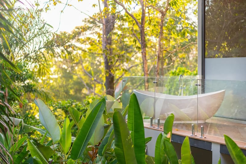 Colíbri Sur · 15. A freestanding bathtub rests on a wooden terrace with a glass railing, offering a view of sunlit tropical foliage.