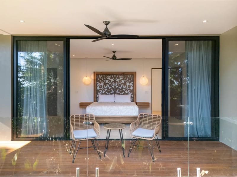 Colíbri Sur · 6. A bedroom with a king-size bed and carved wooden headboard opens through wide glass sliding doors to a private wooden terrace with outdoor seating, overlooking tropical foliage.