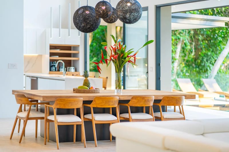 Colíbri Sur · 20. A modern dining space with a large wooden table and seating for eight connects to a contemporary kitchen, opening via glass doors to a sunlit outdoor terrace with tropical views.