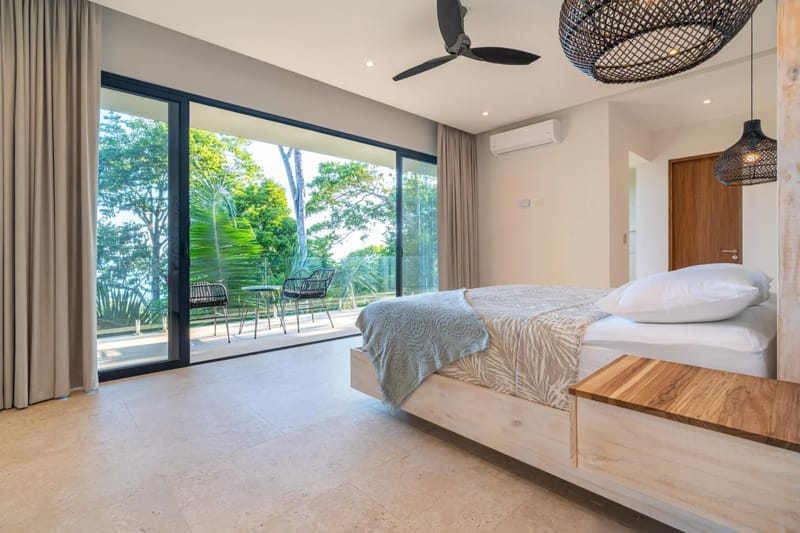 Colíbri Sur · 18. A bright bedroom features a king-size bed, ceiling fan, and air conditioning, with floor-to-ceiling glass doors opening to a private balcony and tropical jungle views.