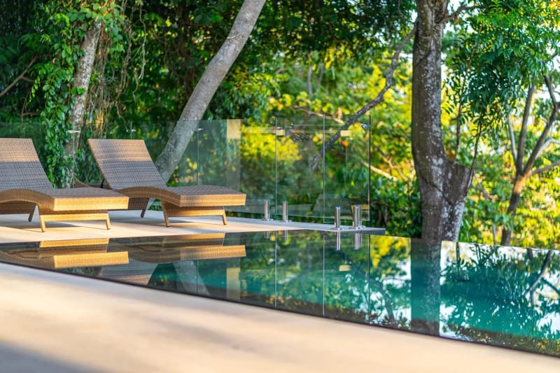 Colíbri Sur · 16. An infinity pool with a clear glass railing provides expansive views of lush tropical foliage, with two lounge chairs positioned on the adjacent terrace in warm natural light.
