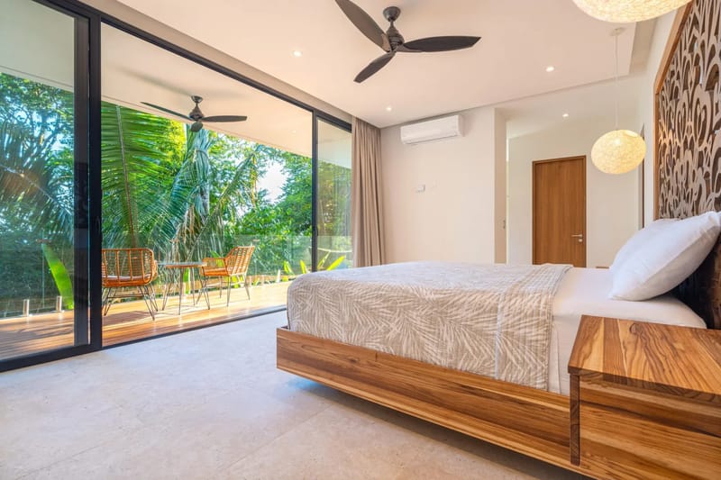 Colíbri Sur · 7. A modern bedroom with a king bed and decorative wood headboard receives natural light through full-height glass doors that open to a private balcony overlooking dense tropical foliage.