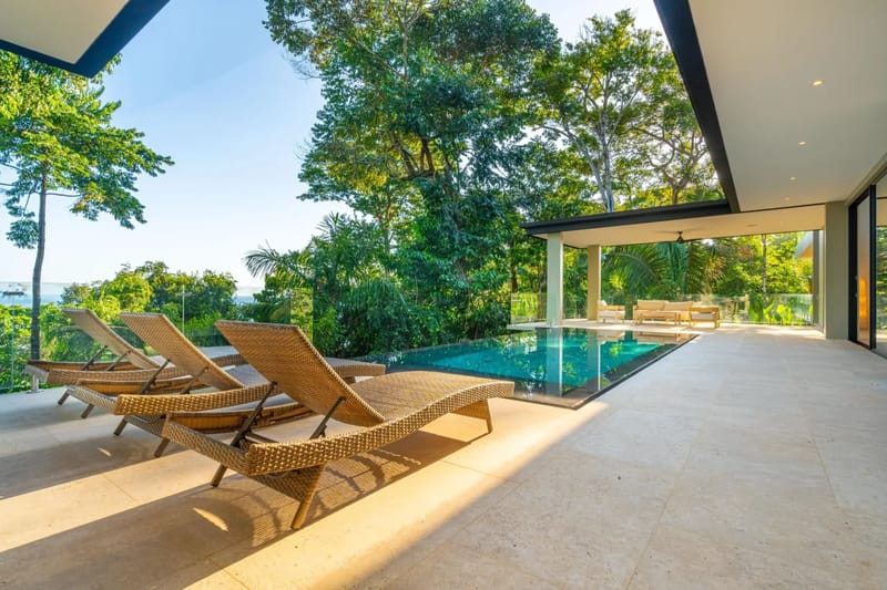 Colíbri Sur · 4. The outdoor area features an infinity pool, a spacious tiled terrace with lounge chairs and a covered seating area, all set against a backdrop of dense tropical foliage and a distant ocean view.