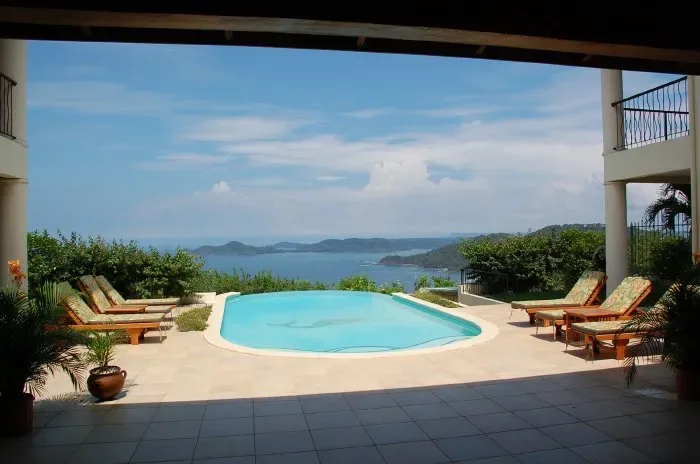 A swimming pool sits on a sunny terrace overlooking the water and distant hills.