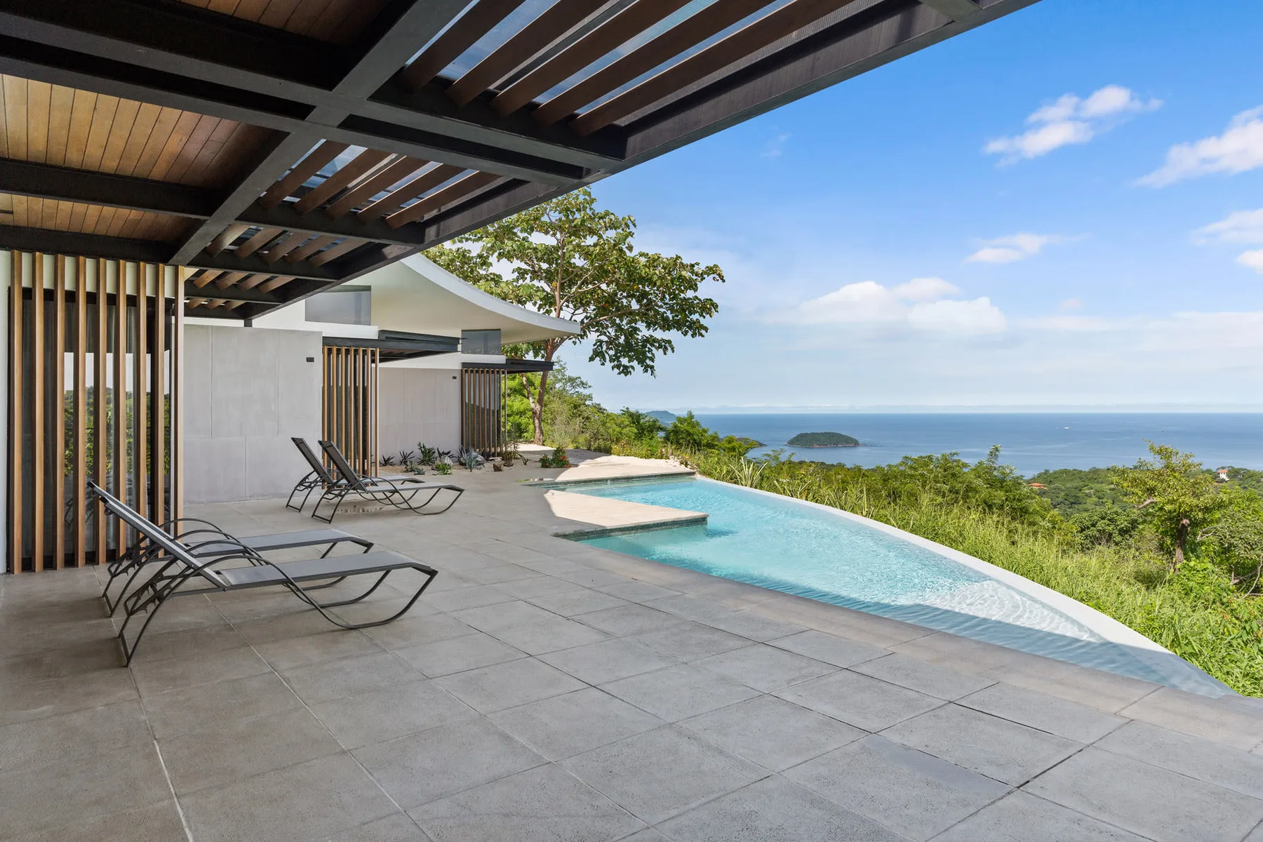 A poolside terrace with lounge seating overlooks the sea beneath a slatted pergola.