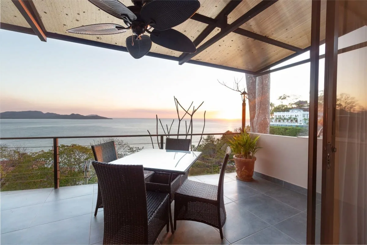 A covered seaside patio with outdoor seating overlooks the water at sunset.