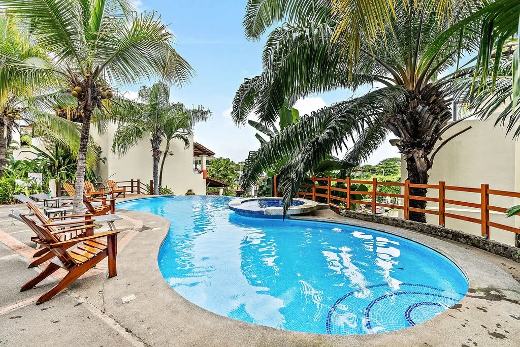 Una piscina exterior curva rodeada de palmeras y asientos para descansar.