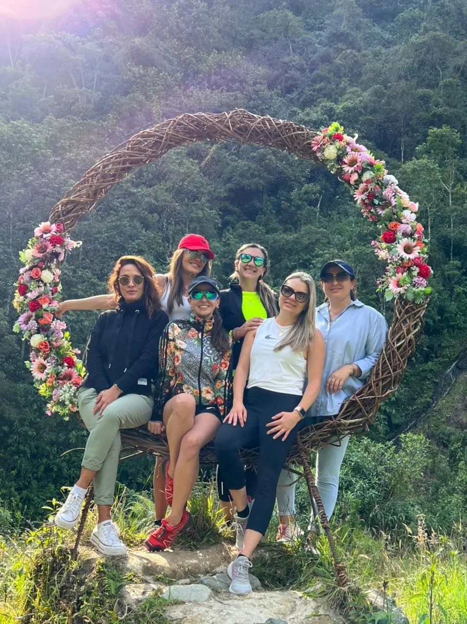A group of people pose on a flower-decorated swing against a forested hillside.