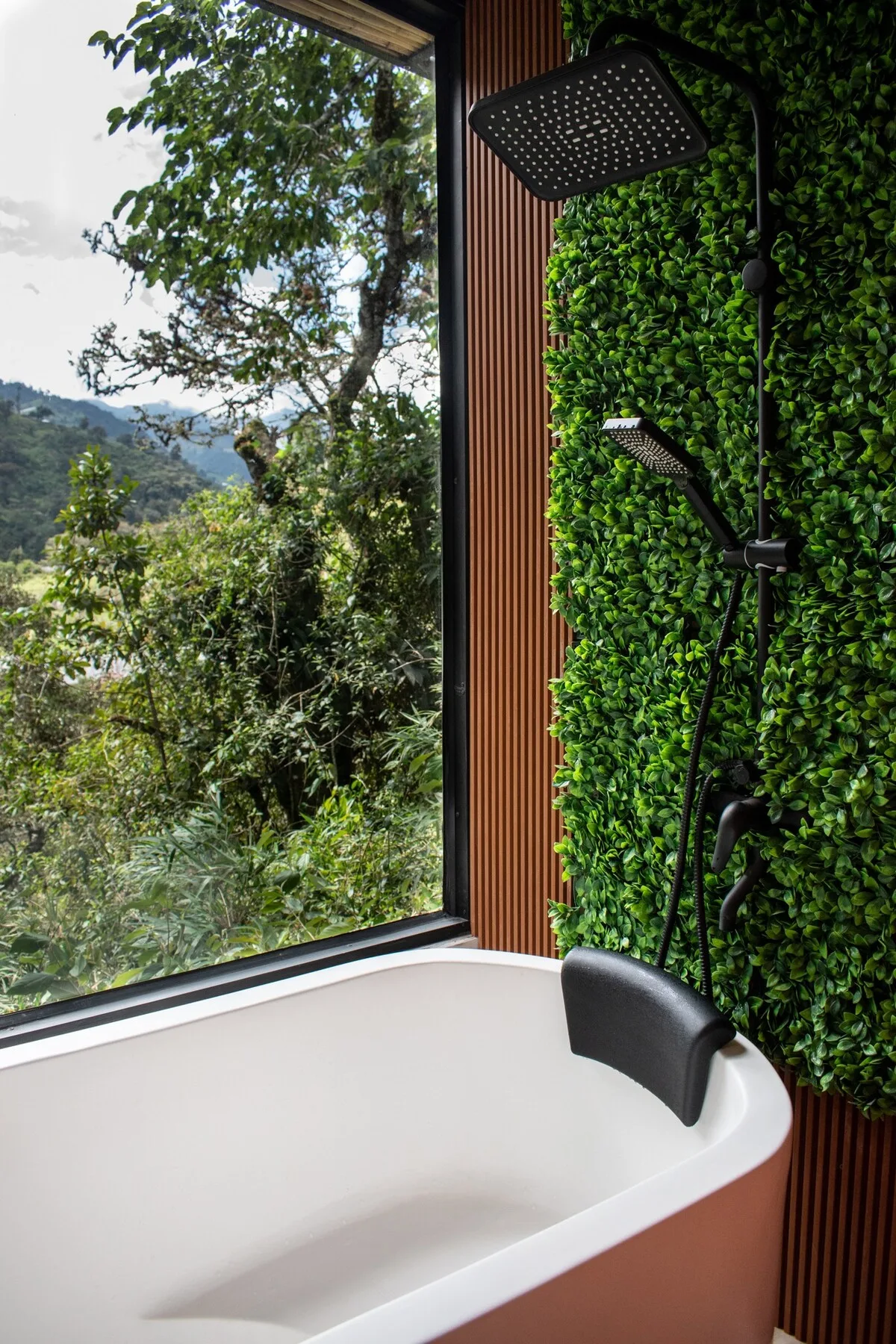 A bathtub sits beside a large window overlooking green hills, with a wall-mounted shower.