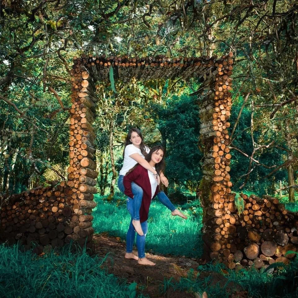 Two people pose beneath a rustic arch made of stacked logs in a wooded setting.