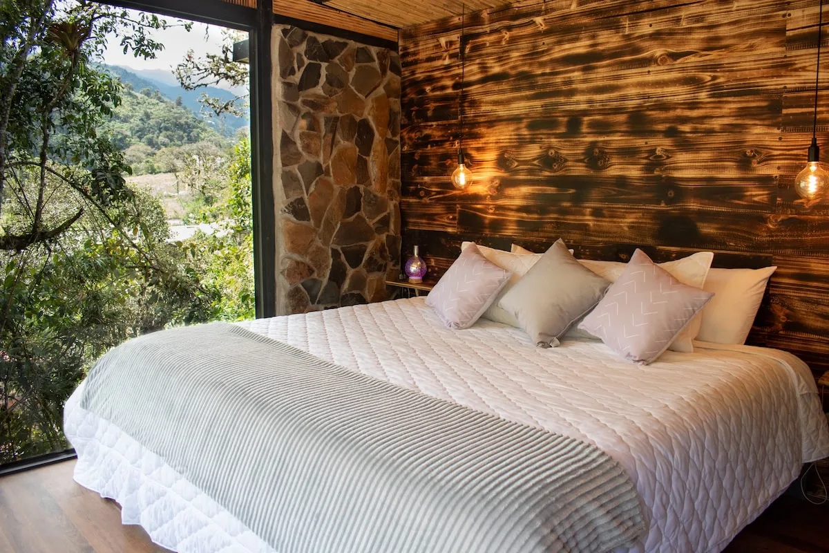 A bedroom with a neatly made bed beside a large window overlooking greenery.