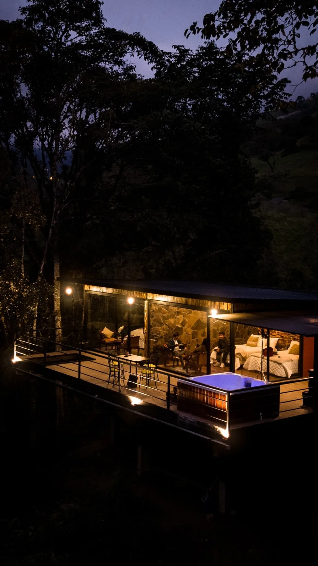 A warmly lit outdoor dining and seating area beneath trees at dusk.