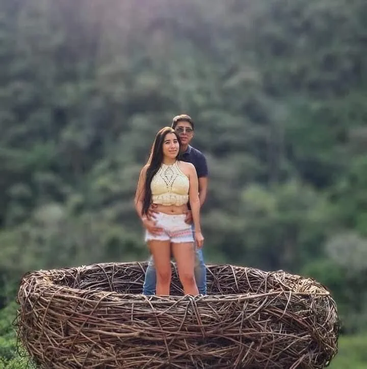 A couple poses inside a large woven nest structure against a wooded hillside.