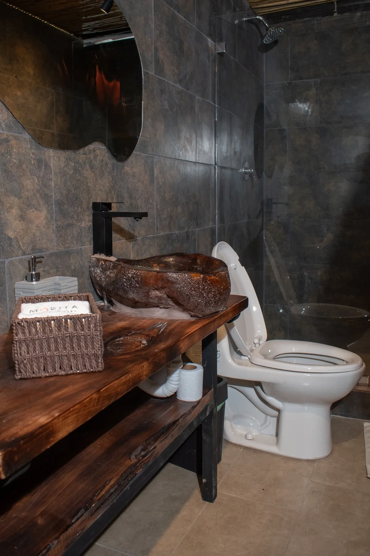 A dark-tiled bathroom with a wooden vanity, vessel sink, and toilet.