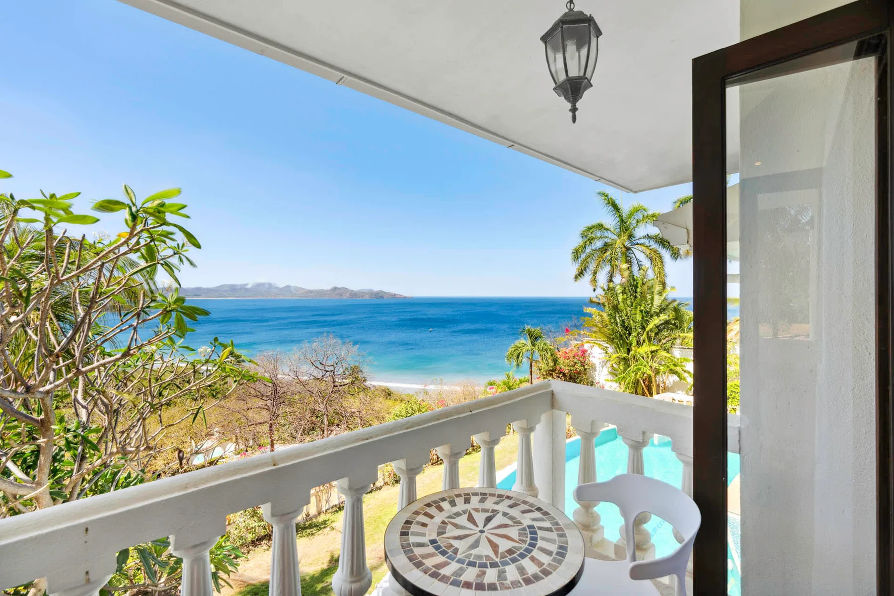 A seaside balcony overlooks the blue ocean, with a small table and seating.