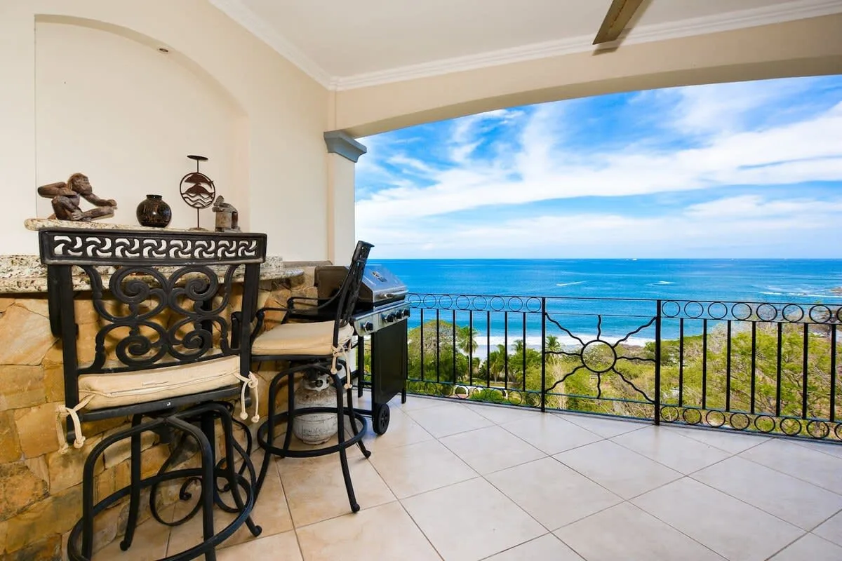 A covered terrace has seating and a grill beside a railing overlooking the ocean.