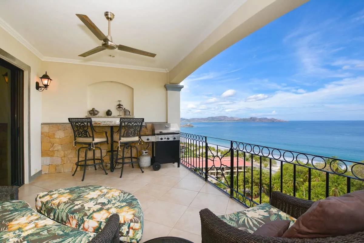 A covered patio has a dining area and seating overlooking the ocean.