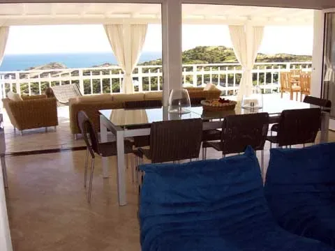 Una terraza cubierta con comedor tiene vistas al mar y a una costa verde.