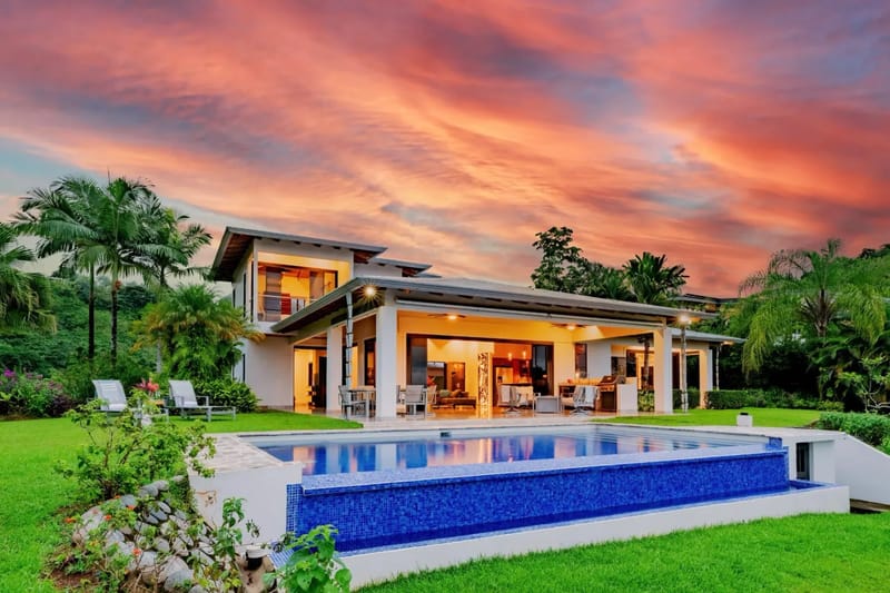 Casa Hacienda Jaguar · 24. The modern multi-level villa features an illuminated open-concept ground floor connecting to a covered porch, overlooking an infinity pool and tropical jungle under a vibrant sunset sky.