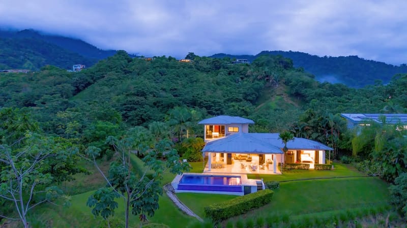 Casa Hacienda Jaguar · 11. The multi-level villa is illuminated at dusk, showcasing an open-concept ground floor with a covered porch and an infinity pool, all set amidst lush, jungle-covered mountains.