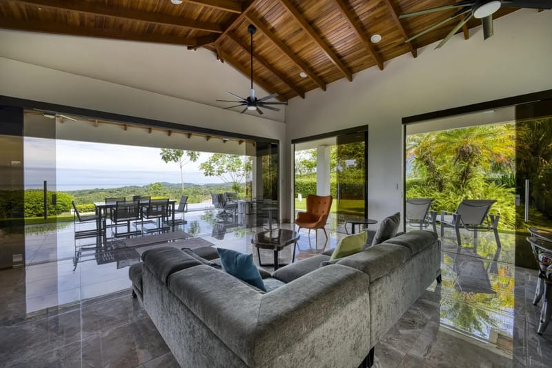 Casa Hacienda Jaguar · 39. An open-concept living space with a vaulted wood ceiling and floor-to-ceiling glass walls connects seamlessly to outdoor dining, offering expansive ocean and jungle views bathed in natural light.