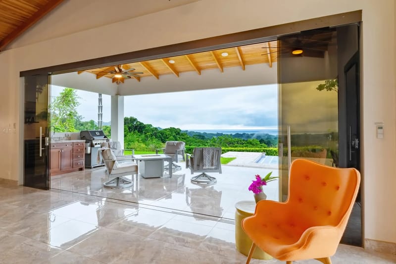 Casa Hacienda Jaguar · 23. Wide sliding glass doors connect the interior to a covered terrace featuring a slatted wood ceiling, outdoor lounge seating, and a barbecue area, all overlooking lush jungle and the distant ocean.