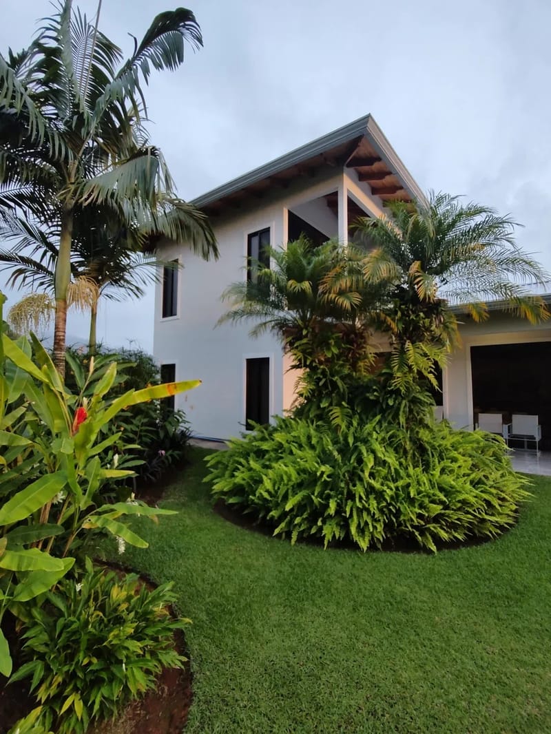 Casa Hacienda Jaguar · 33. A two-story white villa with dark-framed windows and exposed wooden eaves, featuring an upper-level covered balcony and a ground-level covered parking area, surrounded by vibrant tropical landscaping and tall palm trees.