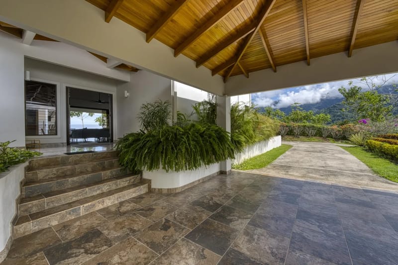 Casa Hacienda Jaguar · 13. This covered villa entrance features a natural wood plank ceiling, tiled steps, and lush tropical plants, with a clear view of the ocean and mountains through the glass doors.