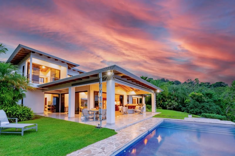 Casa Hacienda Jaguar · 37. The contemporary two-story villa features an open-concept ground floor with glass walls, connecting to a covered outdoor living area and an infinity pool, all set against a backdrop of lush jungle and a vibrant sunset.