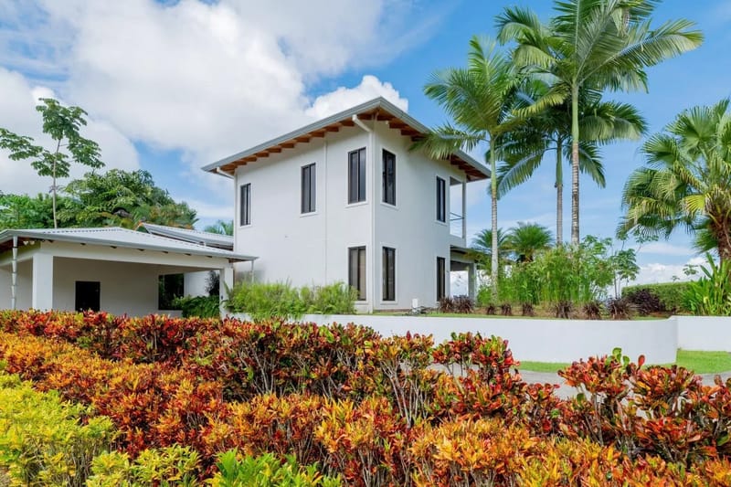 Casa Hacienda Jaguar · 12. A modern, two-story white villa with dark-framed windows, a covered parking area, and an upper-level balcony is nestled within vibrant tropical landscaping and palm trees under a bright blue sky.