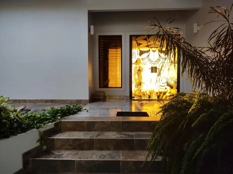 Casa Hacienda Jaguar · 14. The villa's main entrance features a tropical leaf-patterned glass door glowing with warm interior light, flanked by a louvered window, and accessed by tiled steps surrounded by lush exterior plants.