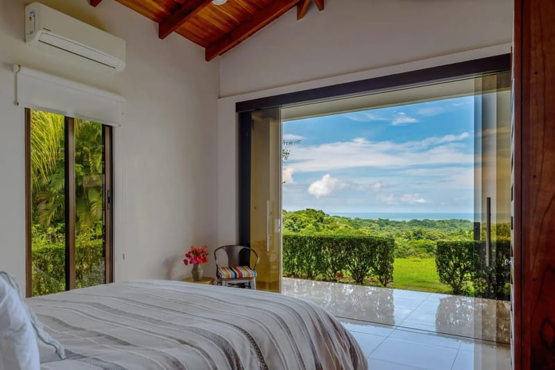 Casa Hacienda Jaguar · 10. A bright bedroom with a queen-size bed and vaulted wooden ceiling opens through wide glass doors to an outdoor area showcasing jungle and distant ocean views.