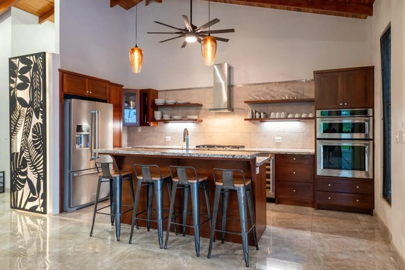 Casa Hacienda Jaguar · 30. This open-concept kitchen features warm wood cabinetry, modern stainless steel appliances, and a granite island with seating under a vaulted ceiling, designed for cooking and socializing while connecting to the villa's outdoor environment.