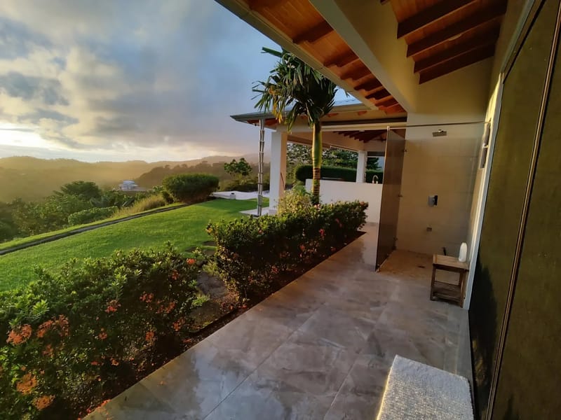 Casa Hacienda Jaguar · 1. A modern tiled outdoor terrace with a rain shower head is covered by a wood-paneled ceiling, overlooking rolling, jungle-covered mountains illuminated by a sunset sky.