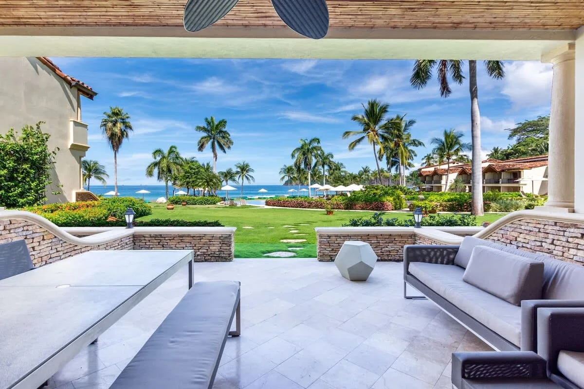 A covered patio with outdoor seating overlooks a palm-lined lawn and ocean.