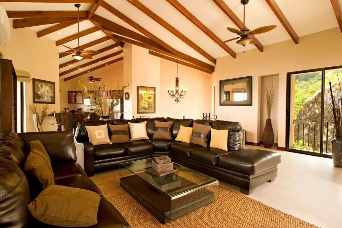 A living room with dark seating, a glass coffee table, and exposed ceiling beams.
