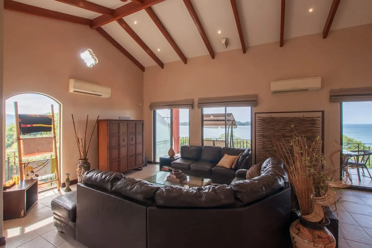 A living room with dark seating, exposed ceiling beams, and glass doors opening onto a balcony.