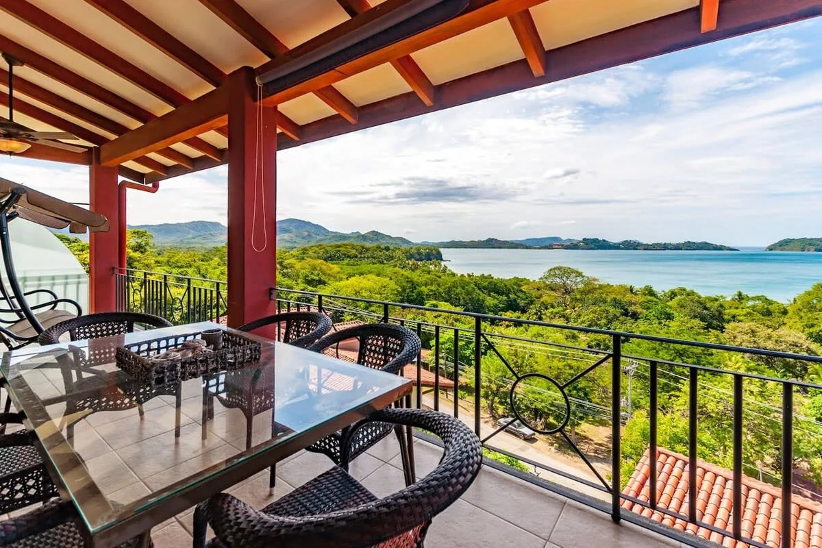 A covered terrace has a dining table and seating overlooking greenery and the water.