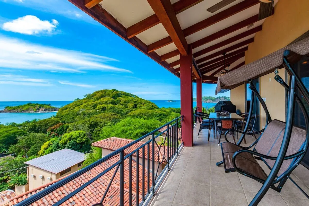 A covered balcony overlooks green hills and the ocean, with seating along the railing.